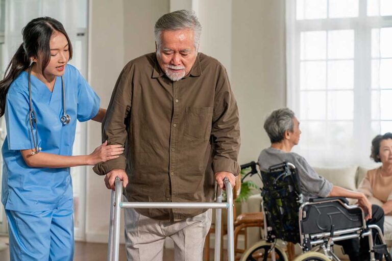 Older adult using a hallway handrail while staff member checks an emergency call system in an assisted living community